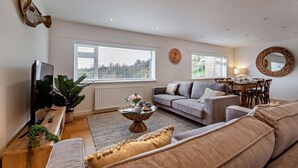 Ein Wohnzimmer mit grauem Sofa, einem Couchtisch mit Blumen, einem Fernseher und einem Essbereich mit Tisch und Stühlen.