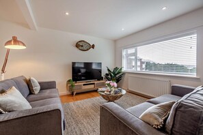 Ein modernes Wohnzimmer mit einem Flachbildfernseher, zwei Sofas, einem Couchtisch mit Dekoration, einer Stehlampe und einem Fenster mit Jalousien.