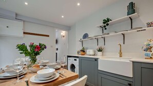 A modern kitchen with a wooden dining table set for a meal, a washing machine, and a built-in oven. - 3 Bed in St. Ives (oc-b29173) (St Ives)