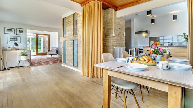 A modern dining area with a wooden table set for breakfast, a brick wall, and a view into the kitchen. - 3 Bed in Walberswick (oc-s29097) (Walberswick)