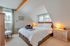 A bedroom with a sloped ceiling, a bed with white bedding, a bedside table with a lamp and alarm clock, and a chair with a view of the outdoors. - 2 Bed in Swanage (oc-k29177) (Swanage)