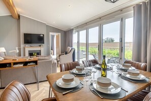 Un espace de repas moderne avec une table dressée pour six personnes, un téléviseur dans le salon et de grandes fenêtres offrant une vue sur l’extérieur.