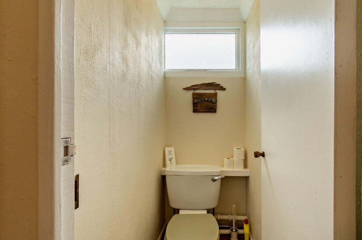 A narrow bathroom with a toilet, a shelf with toilet paper, and a small window above the toilet.