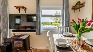 A cozy room with a television, a fireplace, a dining table with a vase of tulips, and a window overlooking a garden. - 2 Bed in St. Dogmaels (oc-s29213) (Saint Dogmaels)