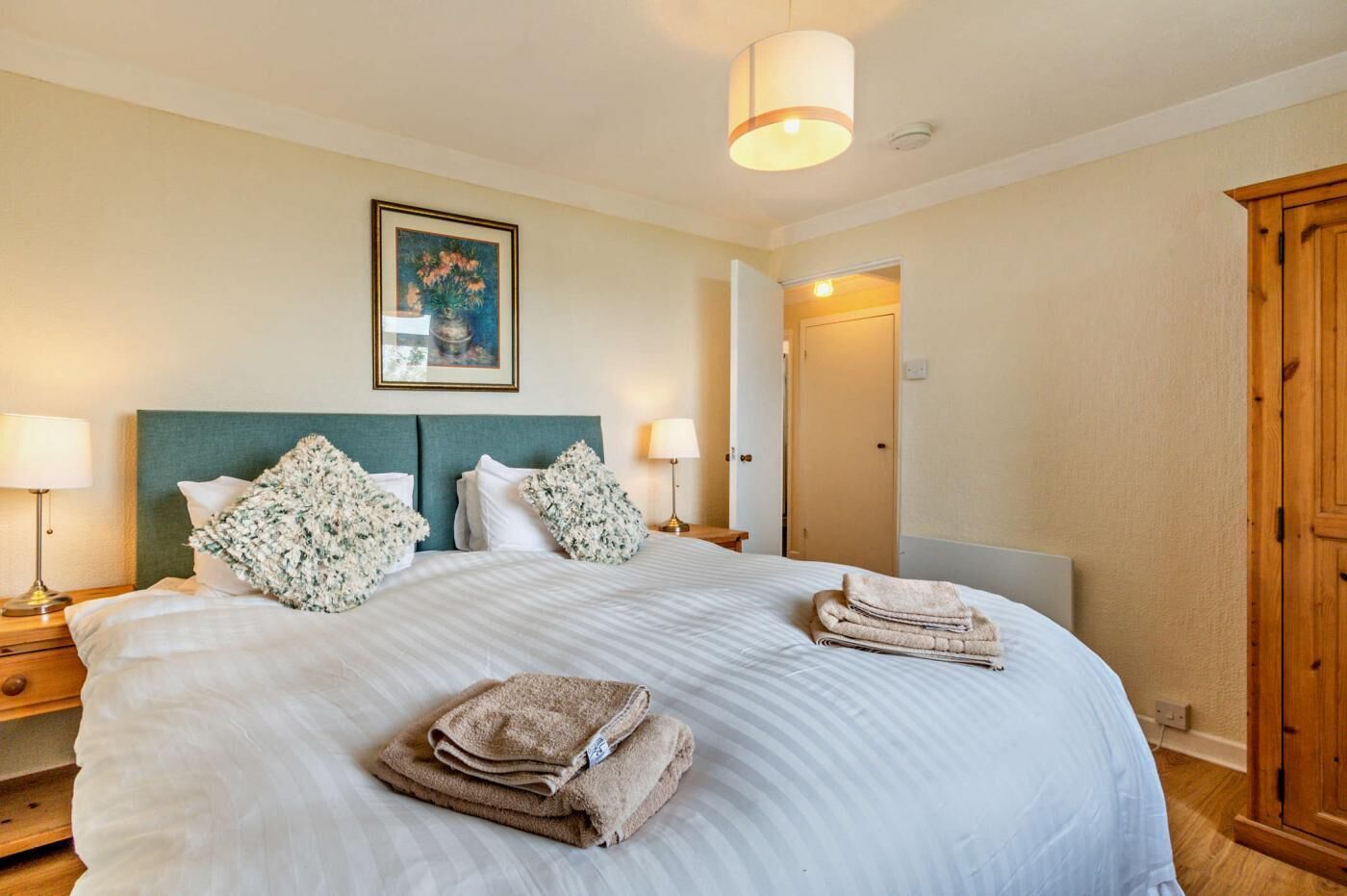 A neatly made bed with white linens and floral pillows, a wooden wardrobe, and a framed artwork on the wall.