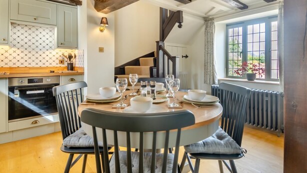 A dining area with a round table set for four, a kitchen with a patterned backsplash, and a view through a window with curtains. - 3 Bed in Weybourne (oc-t29167) (Weybourne)