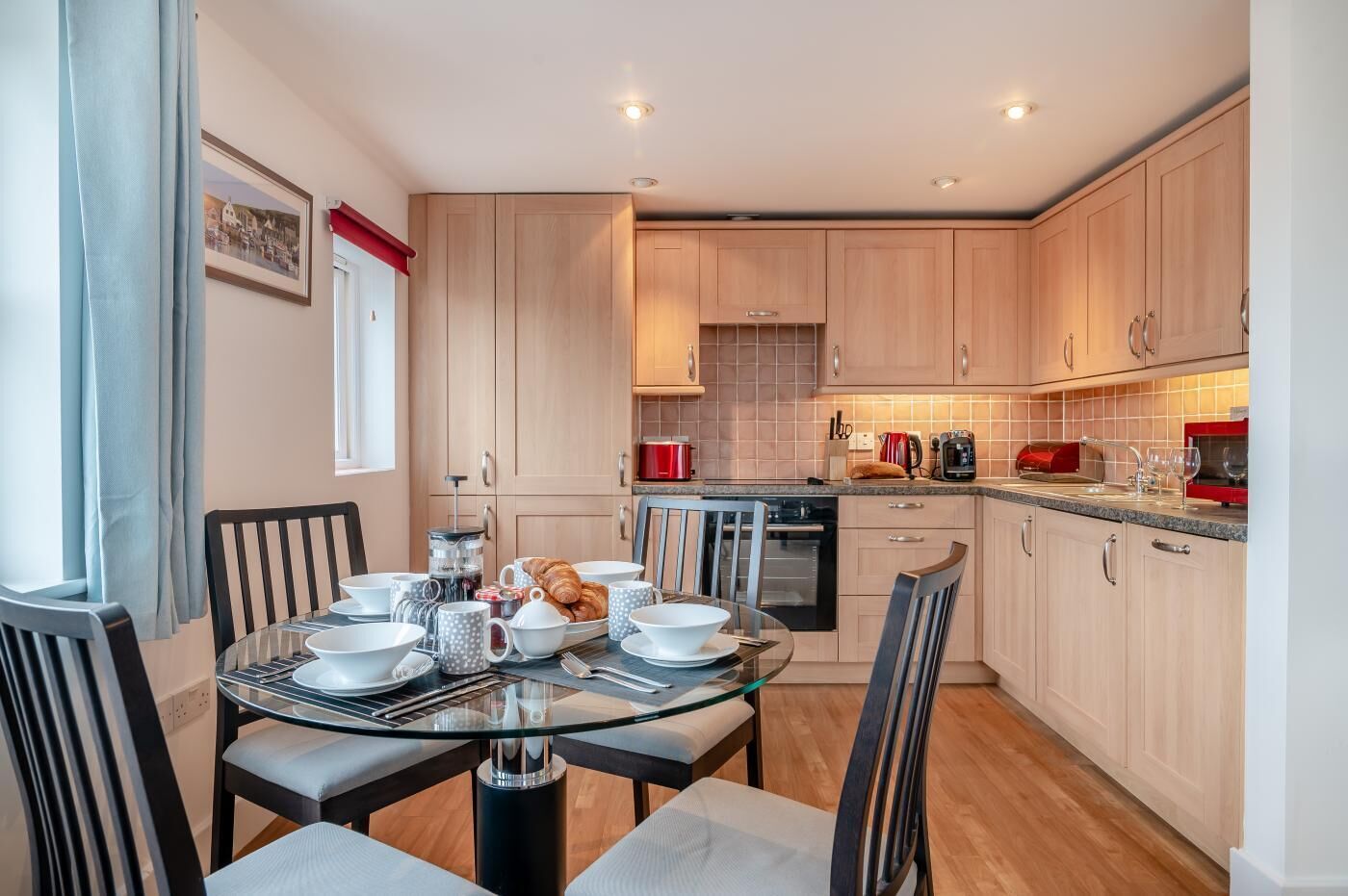A modern kitchen with wooden cabinets, a dining table set with plates, cups, and a croissant.
