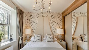 Quarto com uma cama grande, duas mesas de cabeceira com luminárias, um espelho, papel de parede floral e vista para árvores pela janela.