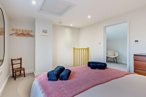 Un dormitorio con cama, dos toallas, una silla de madera, un cabecero de madera, un perchero de pared, un cuadro y una silla blanca en la habitación contigua.