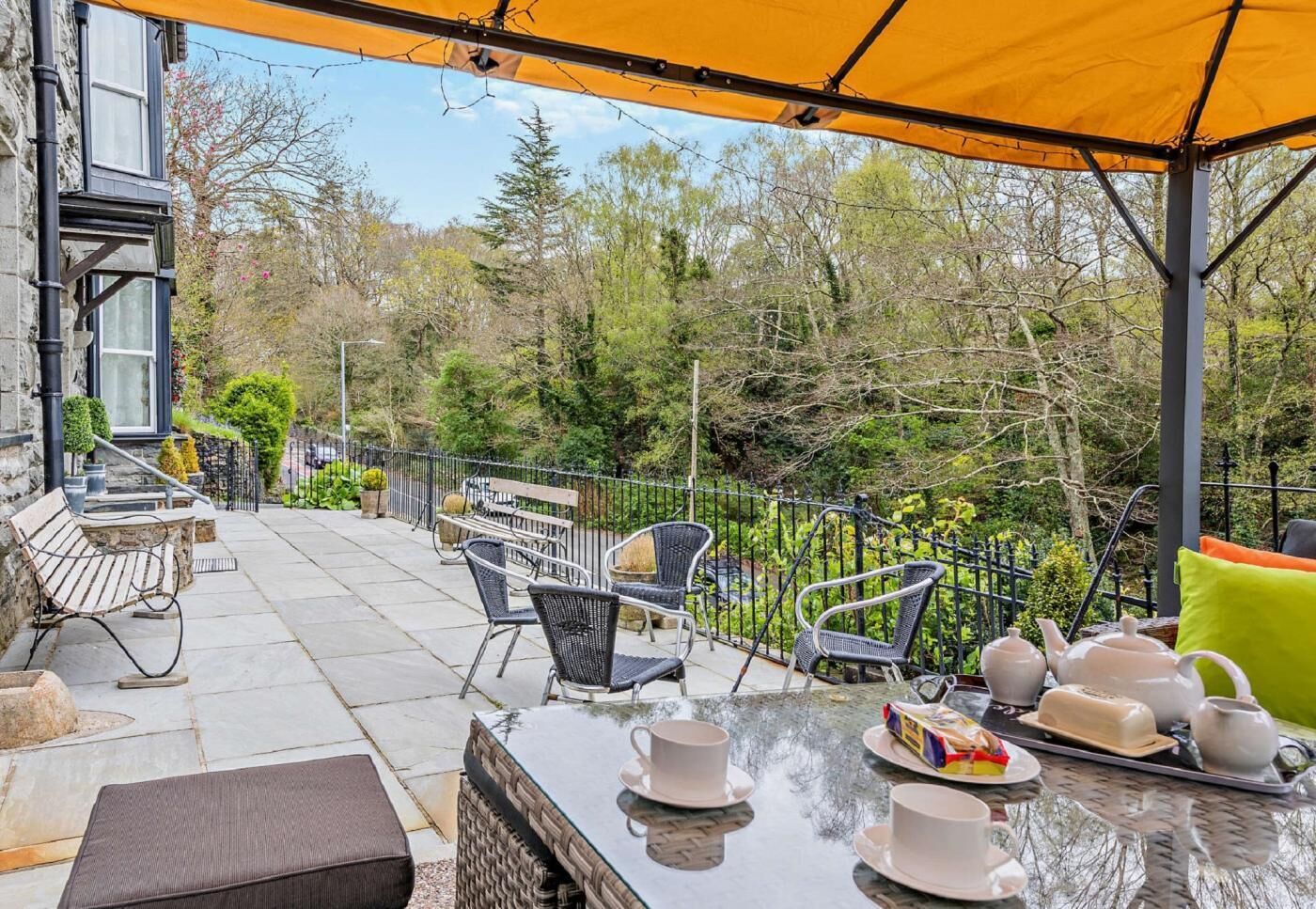 An outdoor patio with a table set for tea, surrounded by chairs and a bench, under a yellow canopy.
