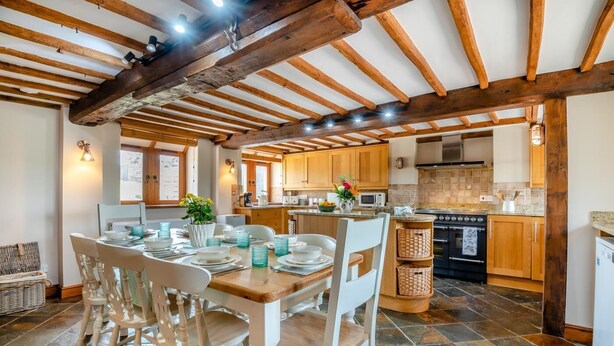 A spacious kitchen with wooden beams, a dining area with white chairs, and a tiled floor. - 4 Bed in Heywood (oc-t29040) (Westbury)