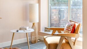 Una habitación acogedora con una mesa redonda, una silla con cojín, una lámpara de pie y una ventana con vistas a edificios.