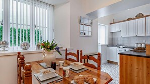 A dining area with a wooden table, chairs, and a vase with flowers. There is a kitchen in the background with white cabinets and a countertop. - 1 Bed in Tenby (oc-p29296) (Tenby)