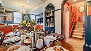 Un coin repas avec une table en bois dressée pour le petit-déjeuner, un lustre et un escalier avec une rampe rouge.