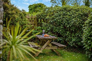 A wooden picnic table with a beverage container and food items, surrounded by lush greenery and a wooden fence. - 2 Bed in St. Issey (oc-t29235) (Saint Issey)
