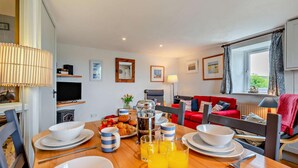 A dining area with a table set for breakfast, including croissants, fruit, and orange juice. There is a red sofa and a television in the background. - 2 Bed in St. Issey (oc-t29235) (Saint Issey)