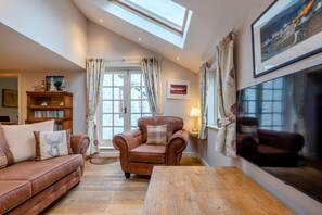 A cozy living room with a brown leather sofa, a wooden coffee table, and a fireplace. There is a bookshelf filled with books and a large window with curtains. - 3 Bed in Freshwater East (oc-w29240) (Freshwater East)