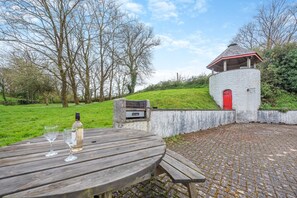 A wooden table with a bottle and glasses, a stone wall, and a red door. - 3 Bed in Freshwater East (oc-w29240) (Freshwater East)