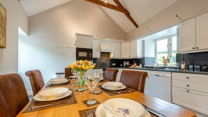 A dining area with a wooden table set for six, surrounded by chairs, with a kitchen in the background. - 3 Bed in Freshwater East (oc-w29240) (Freshwater East)