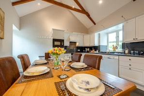 A dining area with a wooden table set for six, surrounded by chairs, with a kitchen in the background. - 3 Bed in Freshwater East (oc-w29240) (Freshwater East)