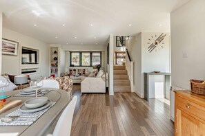 A spacious living area with a sofa set, a dining table with chairs, a staircase, and a kitchen area.