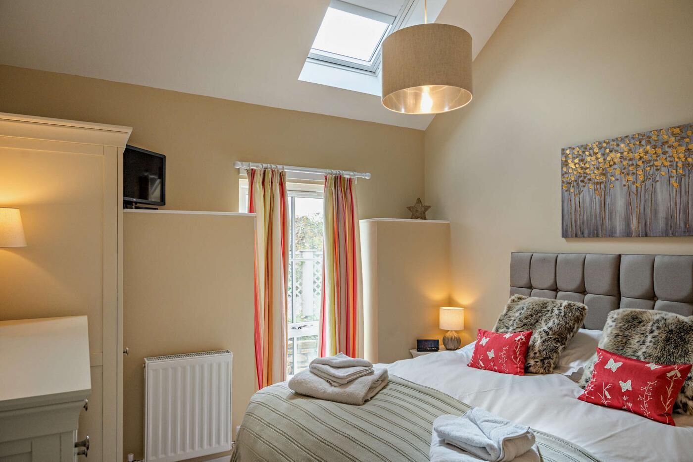 A bedroom with a bed, a radiator, a television, a window with curtains, a ceiling light, and a wall art piece.