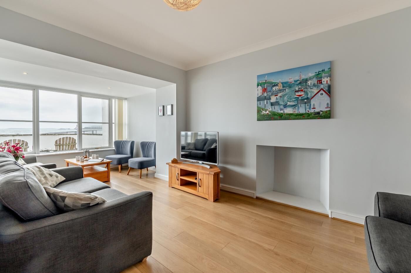 A modern living room with a grey sofa, two armchairs, a coffee table, and a TV on a wooden stand. A painting of a coastal town is on the wall.