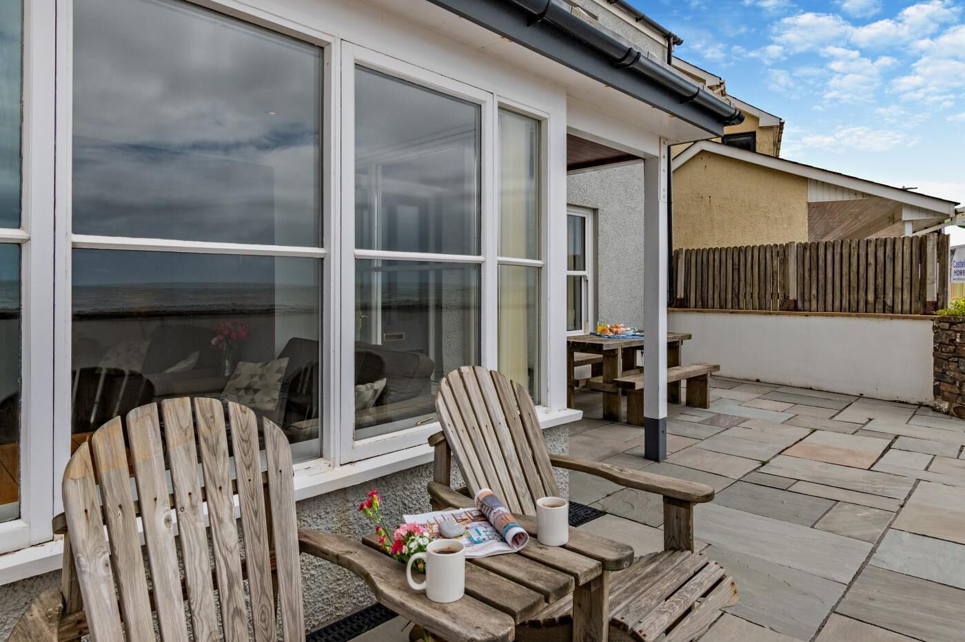 A patio with wooden chairs and a table, a large window, and a stone wall.