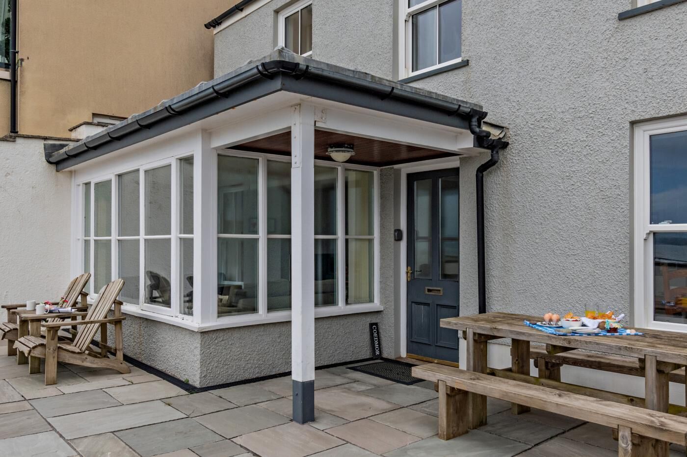 A corner of a building with a bay window, a door, and a small outdoor seating area with a table and chairs.
