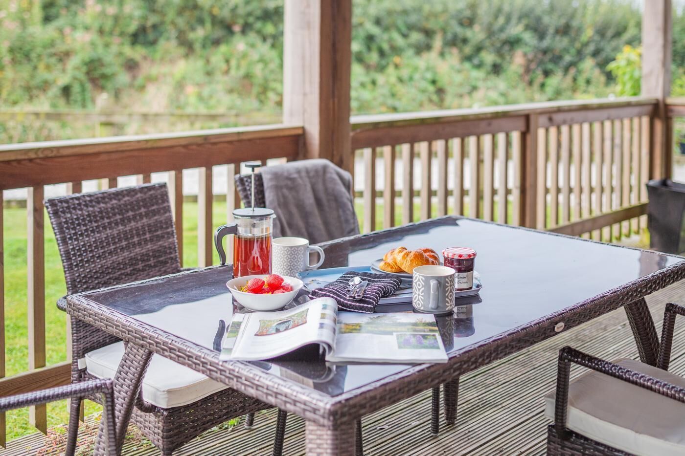 A covered outdoor dining area with wicker furniture, a table with breakfast items, and a view of greenery.