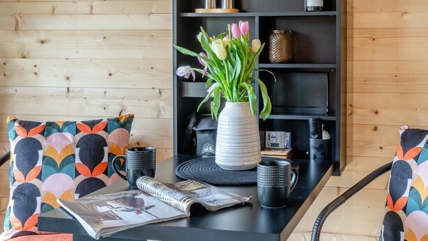 A modern living room with a wooden wall, a black coffee table, two patterned armchairs, a white vase with flowers, and a black bookshelf. - 1 Bed in Cilycwm (oc-t29297) (Cilycwm)
