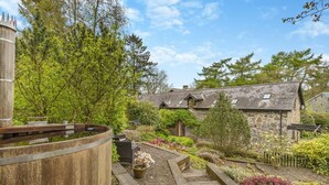 A rustic stone building with a thatched roof, surrounded by a garden with various plants and a wooden barrel structure. - 2 Bed in Knighton (oc-t29330) (Felindre)