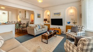A living room with a sofa, coffee table, and TV, a dining area with chairs and a table, and a kitchen with cabinets and a window. - 1 Bed in St. Ives (oc-l29205) (St Ives)