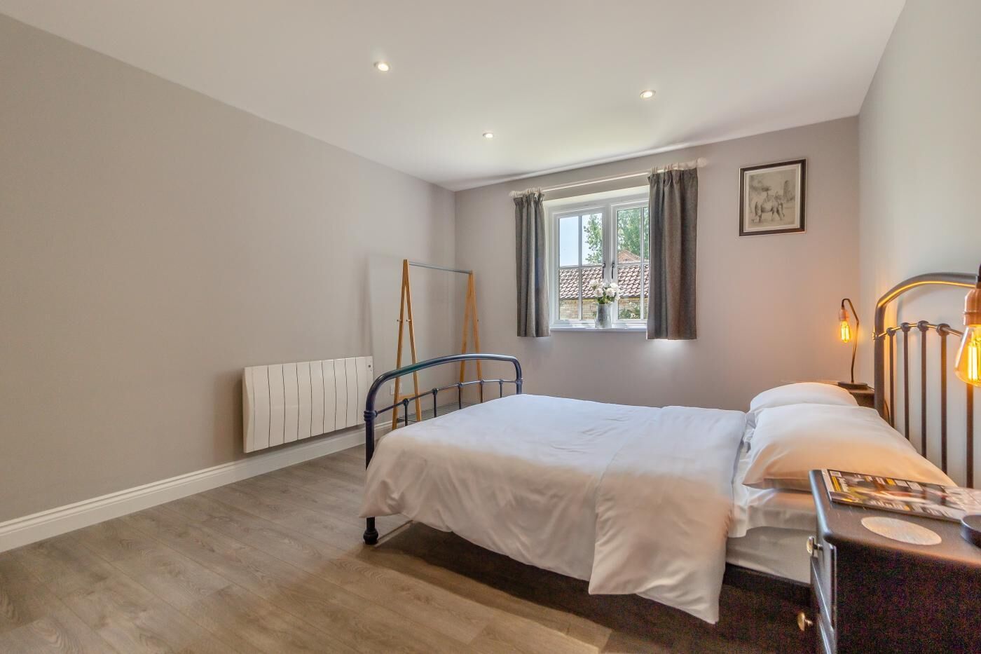 Une chambre à coucher avec un lit, un radiateur, une fenêtre avec des rideaux, une table de chevet avec une lampe et un tableau au mur.