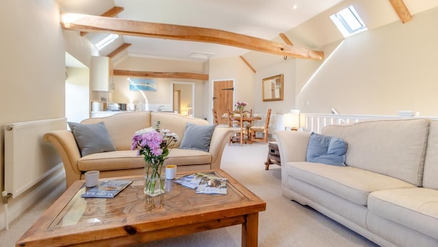 Ein geräumiges Wohnzimmer mit hohen Decken und freiliegenden Holzbalken, ausgestattet mit zwei Sofas, einem Couchtisch und einem Essbereich im Hintergrund.
