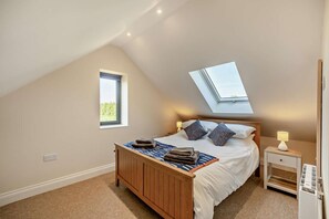 A bedroom with a sloped ceiling, a skylight, a bed with pillows, a bedside table with a lamp, and a power outlet on the wall.