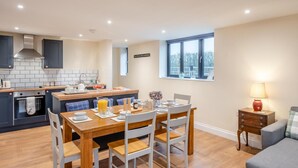 A modern kitchen with a central island, dining table, and chairs. There is a window with a view of trees and a lamp on a bedside table. - 2 Bed in Feering (oc-p29293) (Feering)