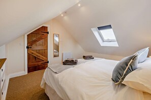Um quarto com teto inclinado, um claraboia, uma cama com almofadas, uma mesinha de cabeceira e uma porta de madeira.
