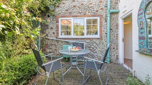 A stone building with a window and a door, a round table with chairs, and a small garden area. - 2 Bed in Holt (oc-m29326) (Holt)