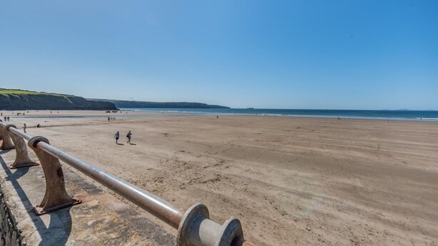 Een strand met een metalen hek in de voorgrond, een zandstrand en een helderblauwe lucht.