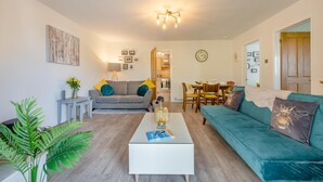 Een moderne woonkamer met een grijze bank, een blauwgroene bank en een witte salontafel. Er is een zithoek met een tafel en stoelen, en een keuken op de achtergrond.