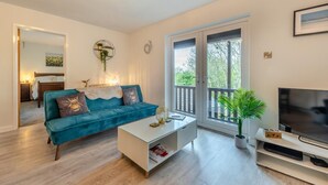 Een moderne woonkamer met een blauwgroene bank, een glazen salontafel, een flatscreen televisie en een balkon met uitzicht op bomen.