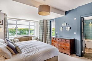 Ein Schlafzimmer mit einem großen Bett, einem Schminktisch, einem Spiegel und Blick auf Grünfläche.