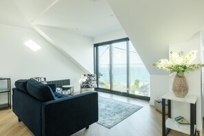 Un salon moderne avec un canapé foncé, une petite table et un fauteuil, une grande fenêtre donnant sur la mer et un vase rempli de fleurs posé sur une étagère blanche.