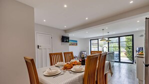 Un comedor con una mesa dispuesta para cuatro personas, sillas de madera, una cocina con un amplio ventanal y una puerta que da a otra habitación.