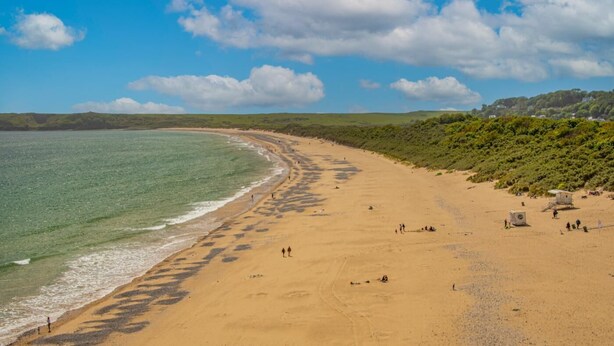 Une vaste plage de sable fin, offrant une vue dégagée sur l’océan et un paysage vallonné en arrière-plan.