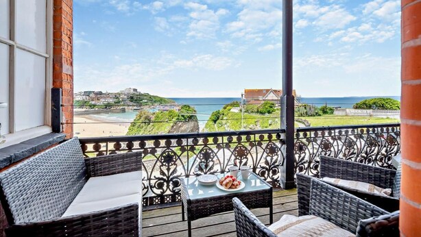Un balcone con mobili in vimini, un tavolo con un piatto di cibo e vista sulla spiaggia e sugli edifici.