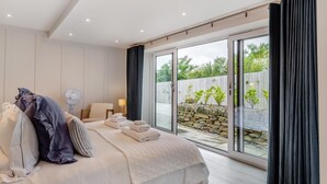 Une chambre avec un grand lit, une table de chevet, une lampe et un ventilateur. Une porte-fenêtre coulissante donne sur un espace extérieur agrémenté d’un mur en pierre et de plantes.