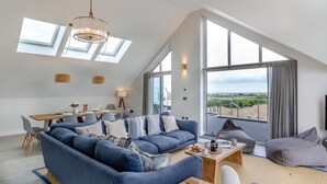 Un salon spacieux avec un grand canapé bleu, une table basse en bois et un coin repas comprenant une table et des chaises. De grandes fenêtres offrent une vue sur la campagne.