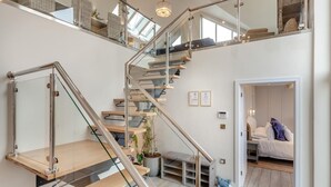 Un escalier moderne avec des rampes en verre et des marches en bois, menant à une chambre à coucher où se trouvent un lit et une plante en pot.